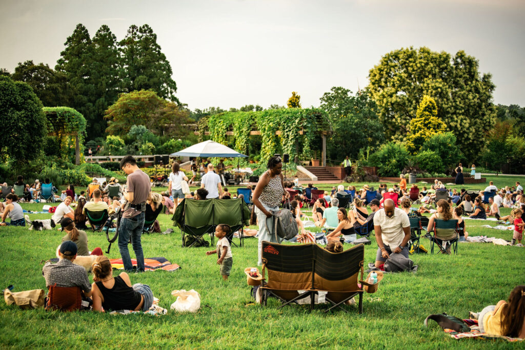 Concert attendees sit in the ellipse meadow while Buck Forty Nine band plays music, with the national herb garden in the background.