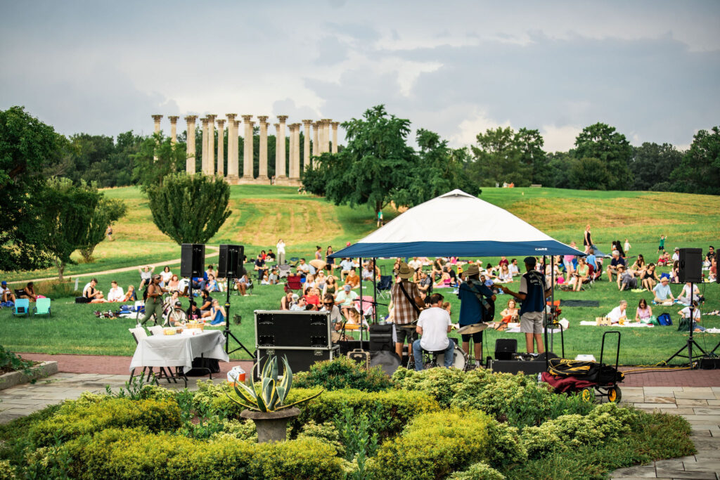 Concert attendees sit in the ellipse meadow while Buck Forty Nine band plays music, with the capitol columns in the background.