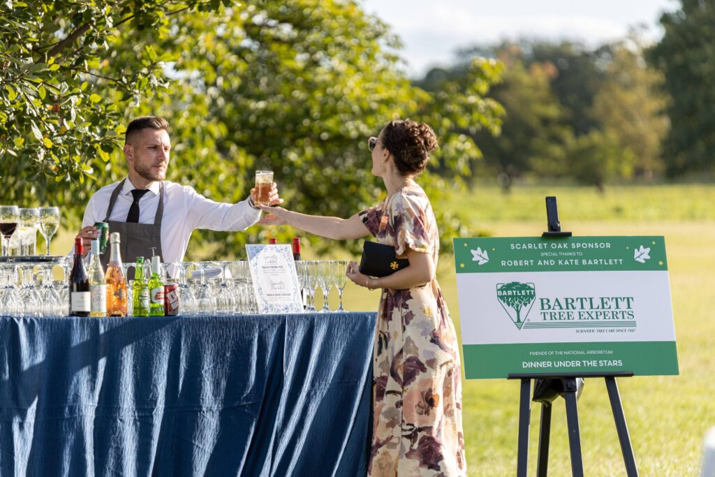 Dinner Under the Stars attendee grabs a drink from a bar during cocktail hour, with a sponsor sign in the background
