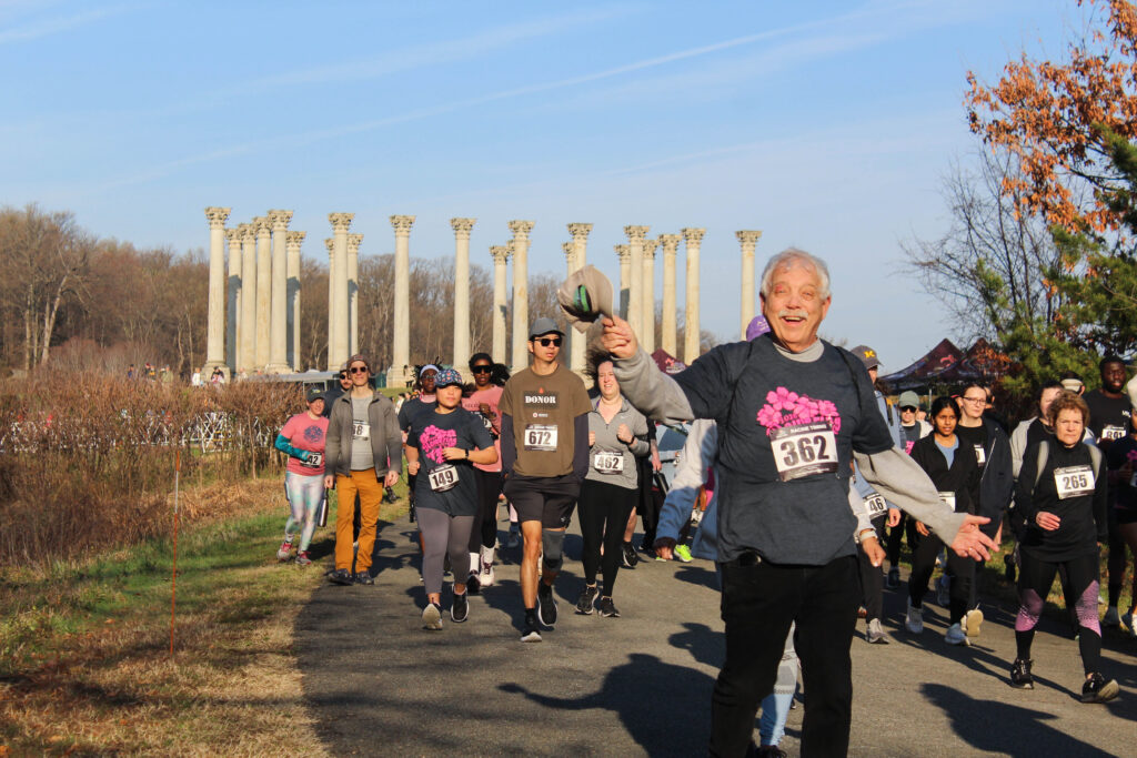 a man smiling and holding out his hat while running the flowering 5k, with the capitol columns in the background