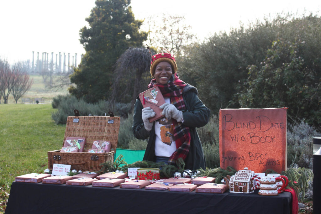 FONA Winter festival vendor wears a scarf, gloves and hat and holds up a book while standing in front of a table.